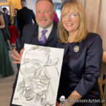 Couple seated holding caricature and laughing, man is bearded, woman has long blonde hair and glasses