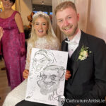 Young newlywed couple seated and laughing, blonde haired bride is wearing a white dress and veil, groom is dark blond and beared, wearing a dark suit with a white bow-tie. They are holding a black and white head and shoulder caricature of themselves, surrounded by lovehearts. The caricature is drawn by Allan Cavanagh