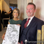 Seated couple laughing holding black and white caricature of themselves drawn by Allan Cavanagh