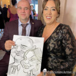 Man in purple suit jacket and woman in black dress laughing holding a caricature by Allan Cavanagh