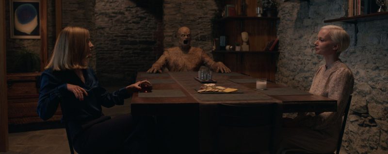 Scene from Oddity Film 2024. Two women sitting at a large dining table in a dimly-lit room. At the head of the table sits a wooden sculpture of a figure with hands resting on the table and an expression of horrific silent roar on its face.